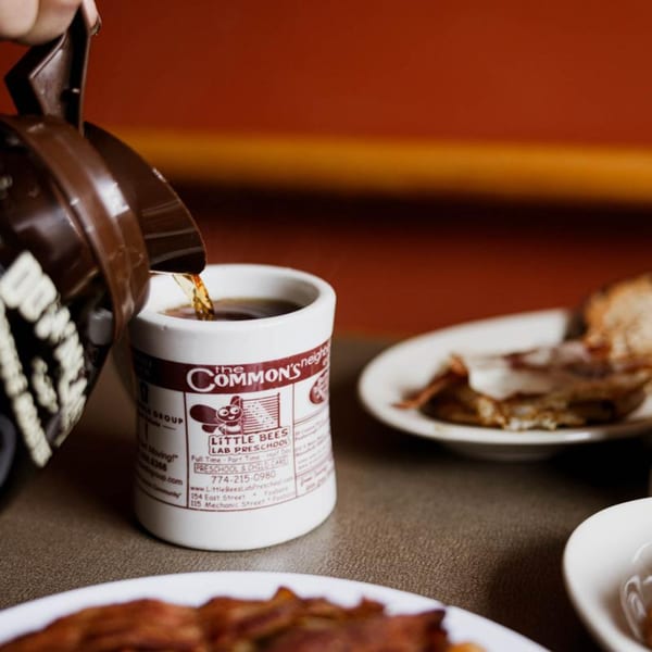 Coffee pouring into a mug with The Commons logo, alongside breakfast plates.