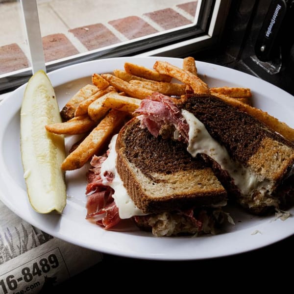 Reuben sandwich with fries and a pickle spear on a white plate, by a window.