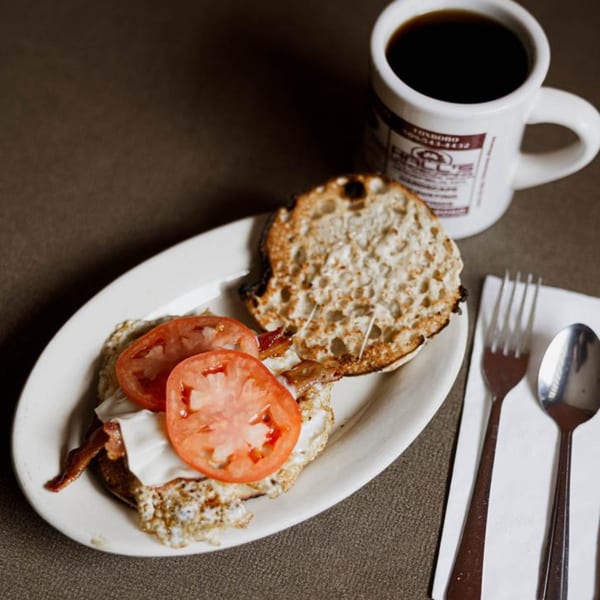 Breakfast sandwich with bacon, egg, cheese, and tomato on an English muffin, with a coffee.