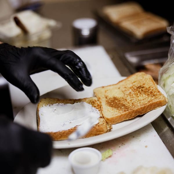 A gloved hand spreads cream cheese on toasted bread, preparing a breakfast item at The Commons.